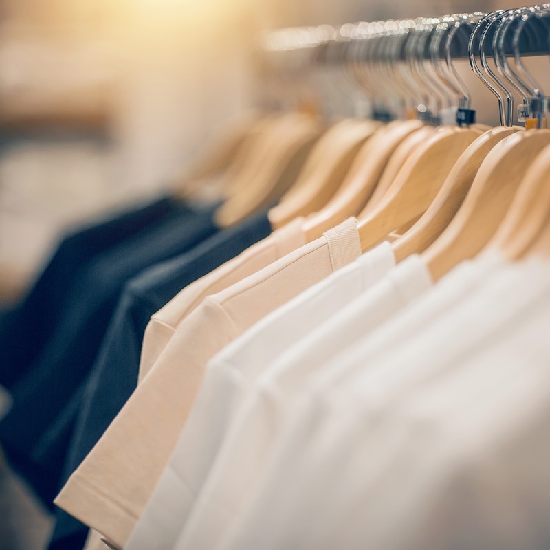 Vêtements Basiques en Rangée : T-shirts sur Cintres T-shirts unis beiges et blancs suspendus sur des cintres en bois sur une tringle métallique, éclairage doux et chaud.