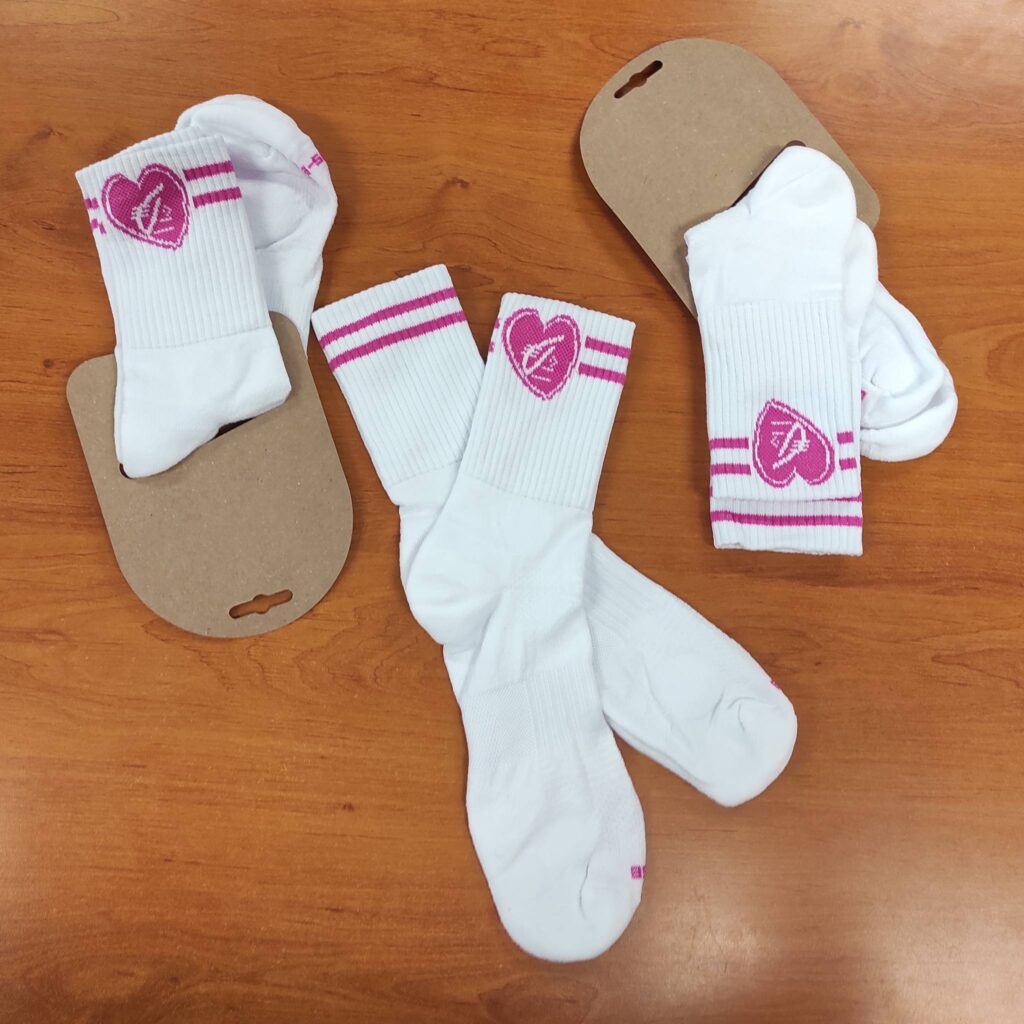 Trois paires de chaussettes blanches, logo cœur et rayures roses vifs, présentées avec leurs étiquettes en carton kraft sur un fond bois.
