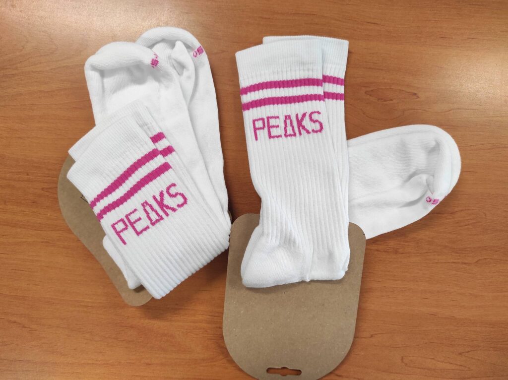 Chaussettes blanches côtelées PEAKS avec bandes magenta, présentées sur un emballage carton découpé, sur une surface en bois.