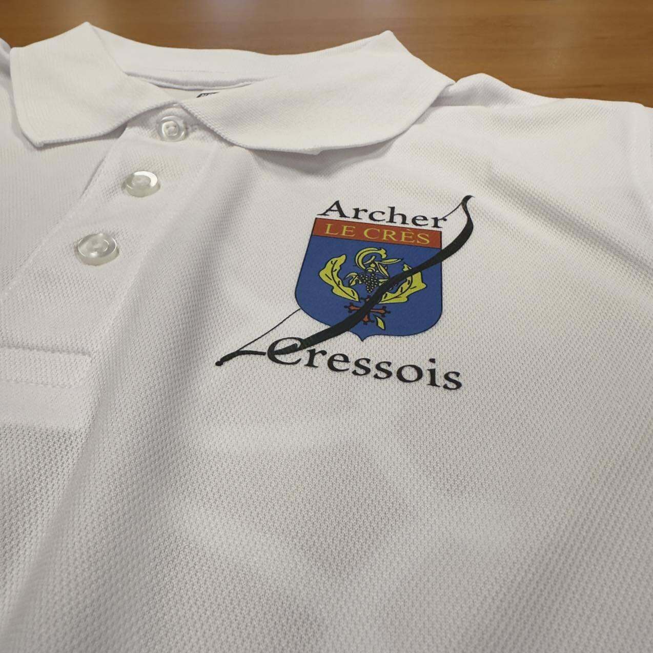 Polo d'archer blanc, tissu piqué, portant le logo du club : "Archer LE CRÈS". Blason bleu traversé par un arc noir, surmontant "Cressois".