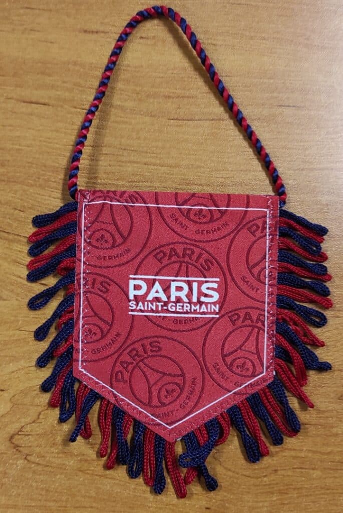 Fanion PSG rouge avec franges bleues et rouges, logo Paris Saint-Germain et cordelette de suspension.