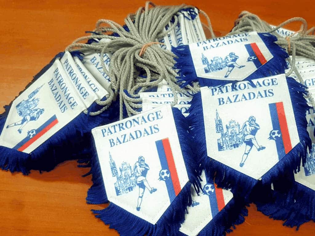 Fanions du Patronage Bazadais avec logo de footballeur et cathédrale, bordés de franges bleues et cordons gris.