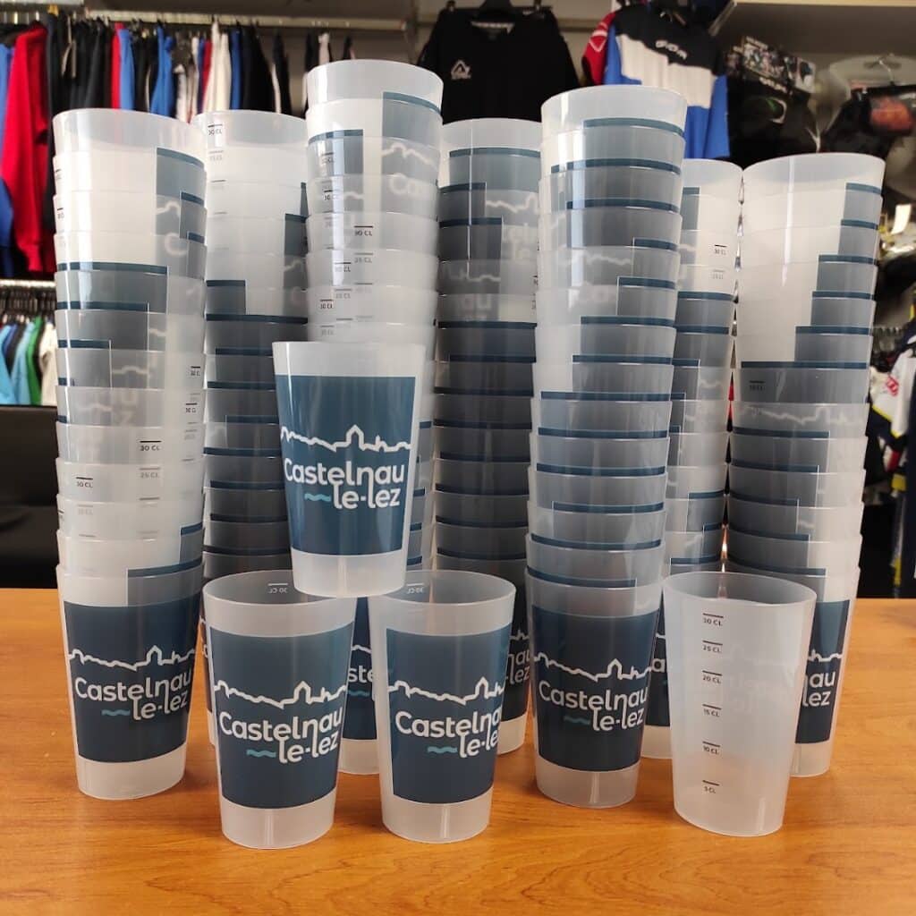 Piles de gobelets réutilisables avec logo Castelnau-le-Lez et graduations, posés sur un comptoir en bois.