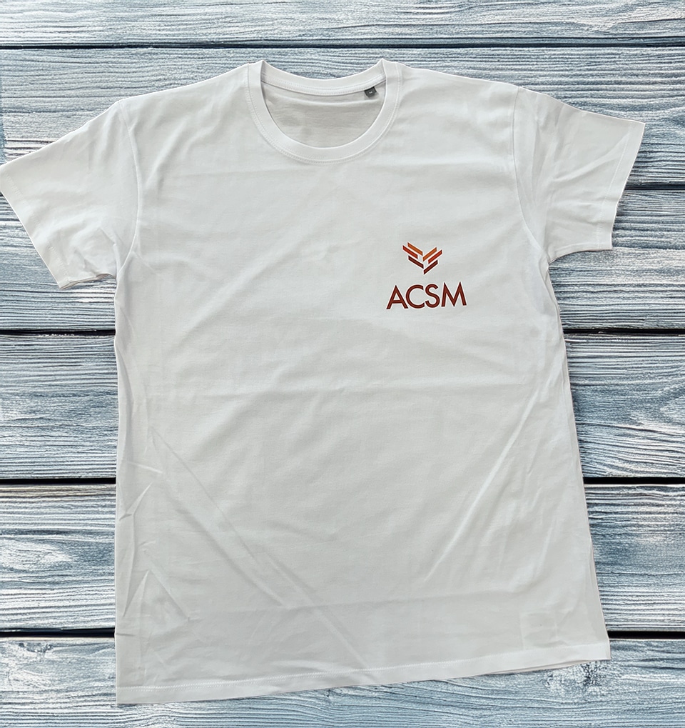 T-shirt blanc avec logo ACSM et emblème géométrique orange posé sur un fond de planches en bois bleu.