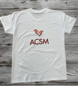 T-shirt blanc avec logo géométrique orange et texte ACSM marron, posé sur un fond en bois gris.