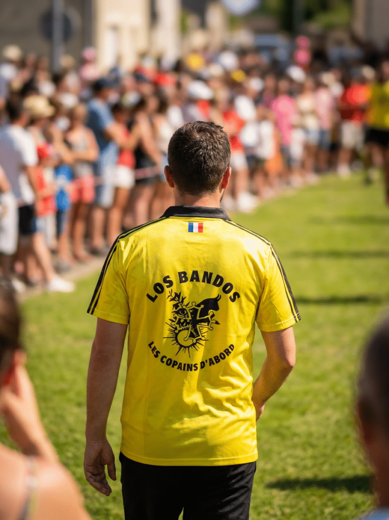 Los Bandos Les Copains d’Abord : Maillot jaune et fête Homme de dos en maillot jaune Los Bandos marchant vers une foule lors d'une fête locale ensoleillée.
